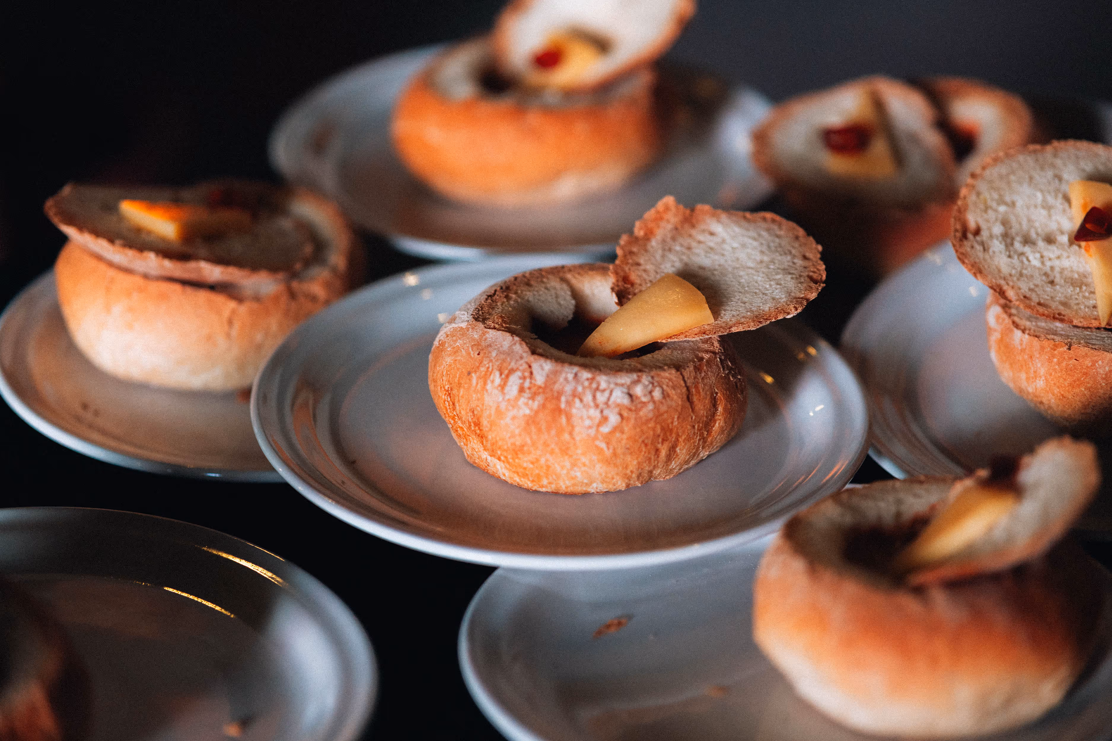 Several bread bowls with lids, each containing a piece of cheese and a red garnish, served on white plates.