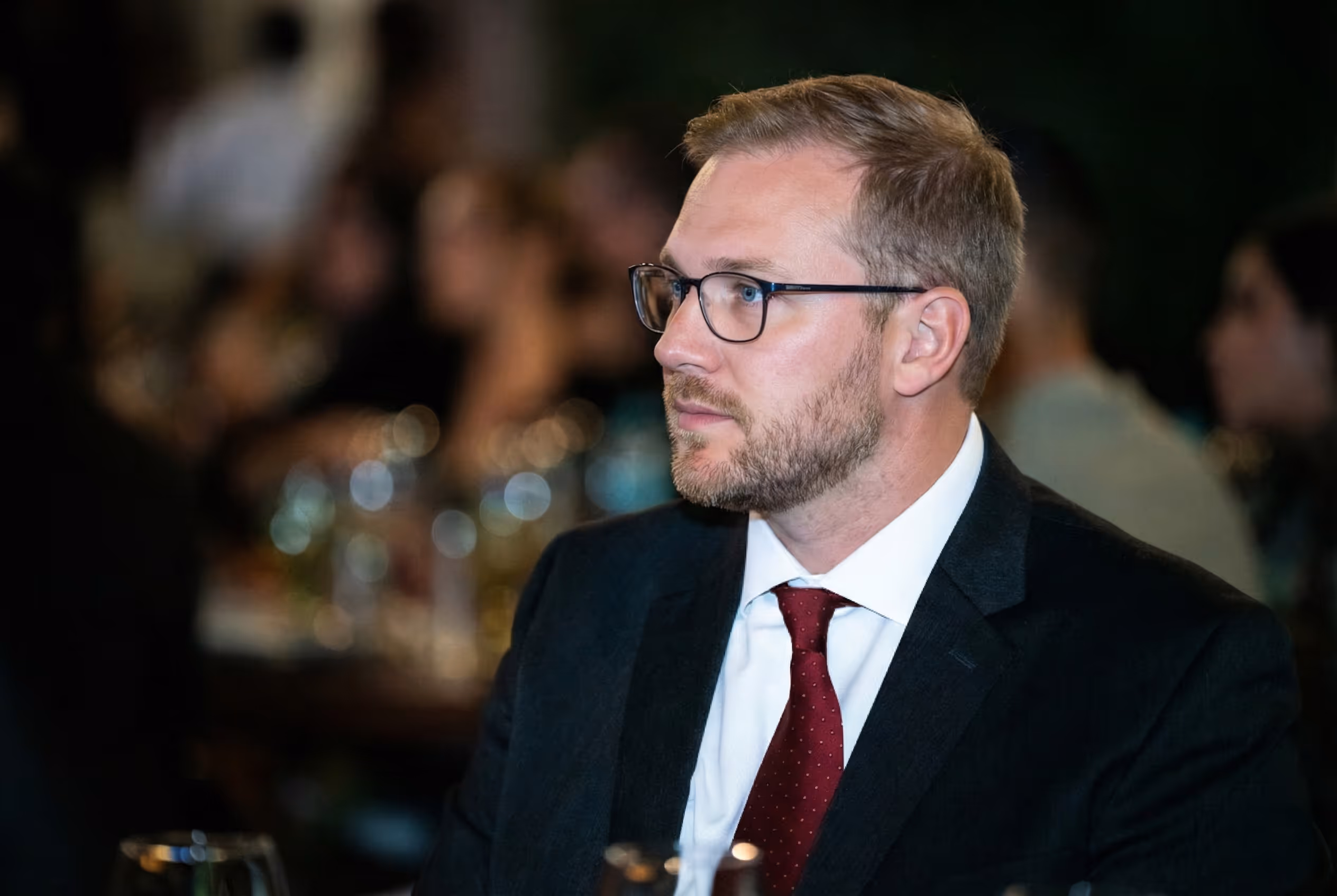 Man with glasses and beard wearing a dark suit and red tie looking to the side at an event.