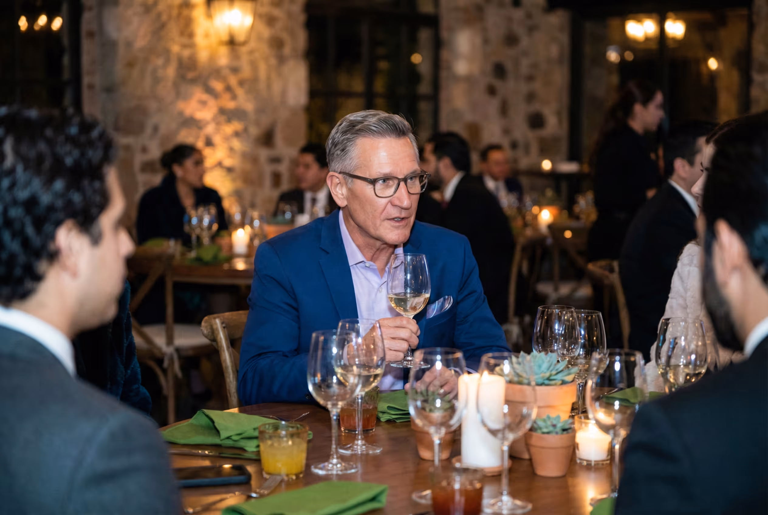 Man in blue suit holding a glass of white wine while seated at a dinner table with multiple glasses, candles, and small potted plants.