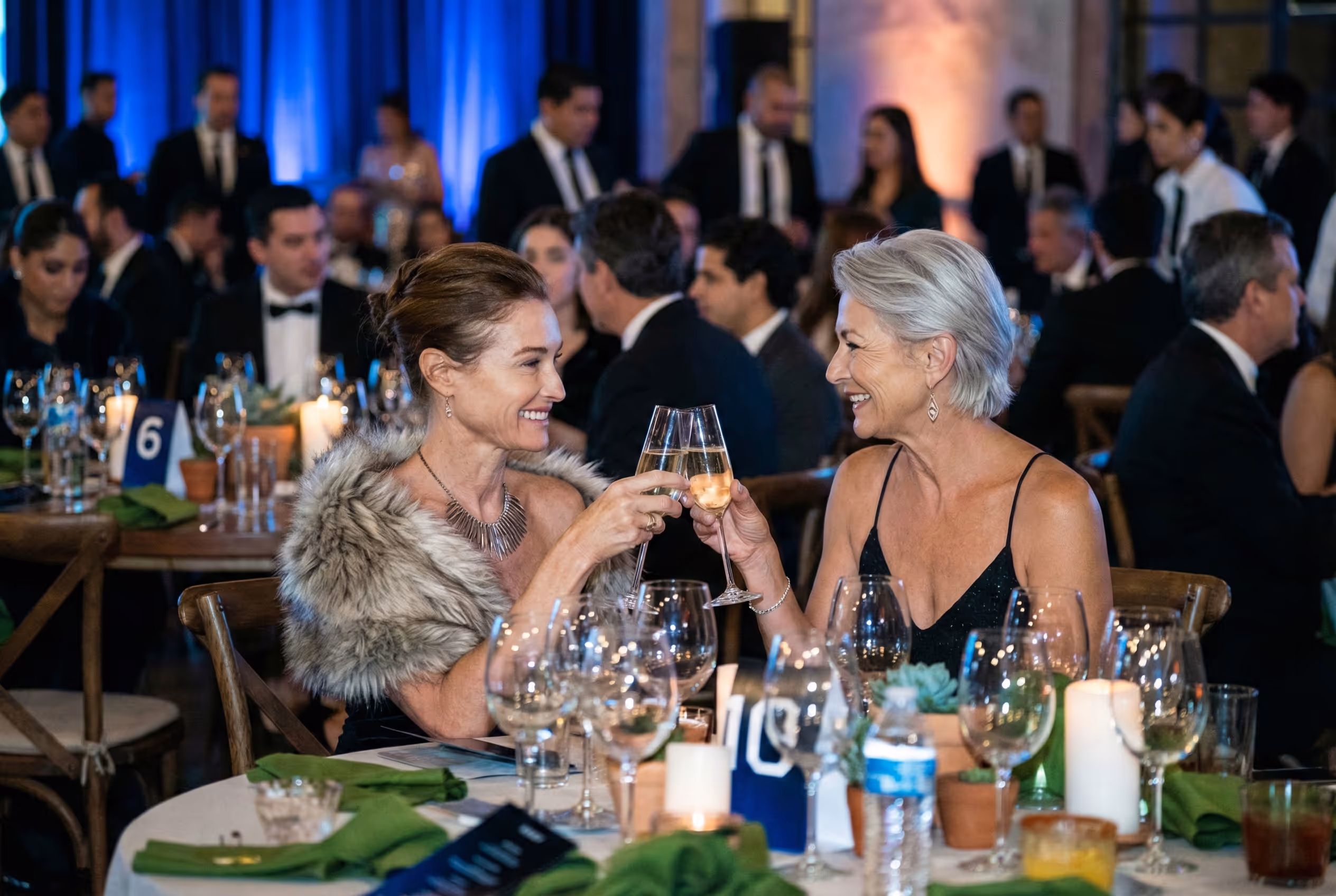 Two elegantly dressed women smiling and clinking champagne glasses at a formal dinner event.