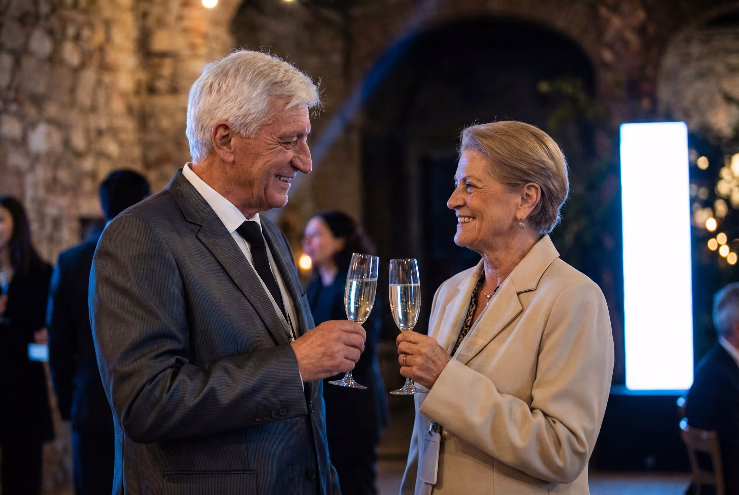 Elderly man and woman smiling and clinking champagne glasses at a formal event.
