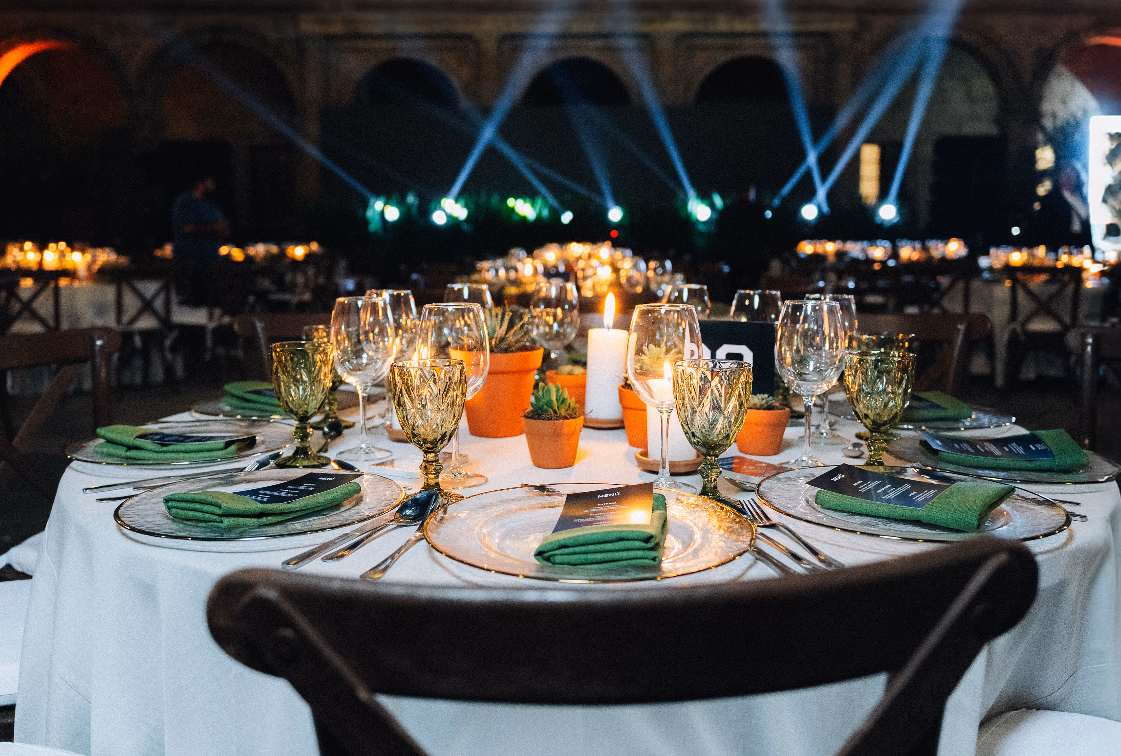 Elegantly set round dining table with white tablecloth, green napkins, glassware, potted succulents, and a lit candle in a dimly lit event space.