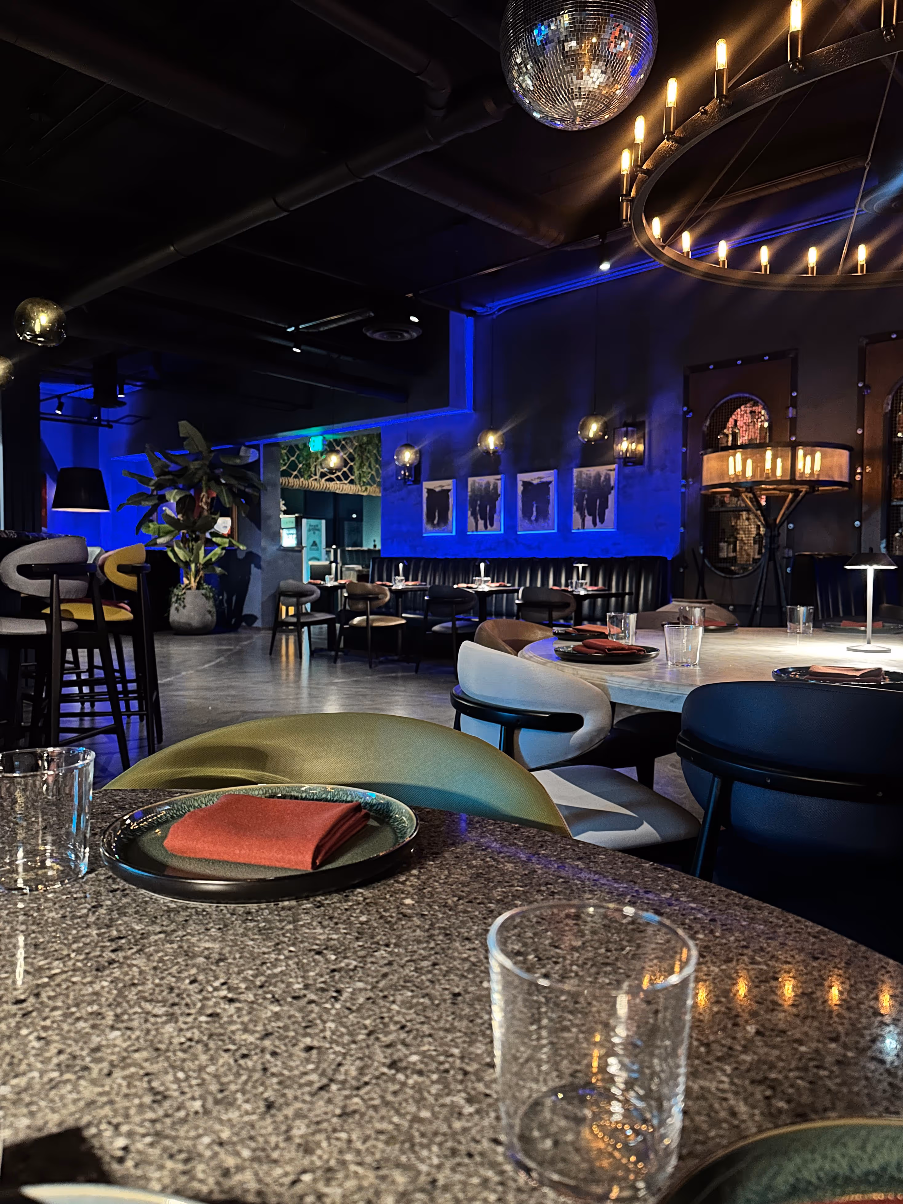 Modern restaurant interior with round tables set with glasses and napkins, blue ambient lighting, and a disco ball hanging from the ceiling.