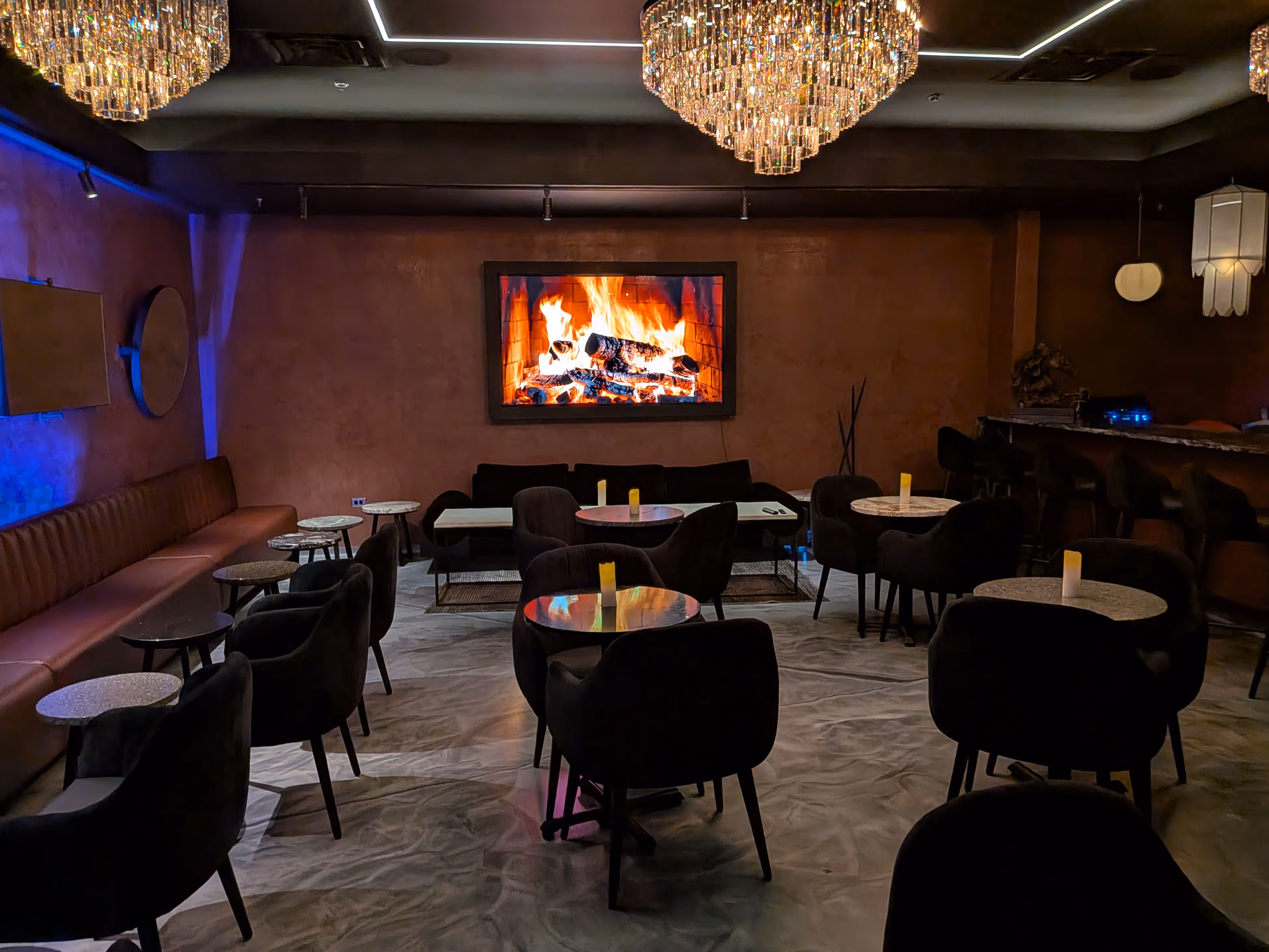 Dimly lit lounge with round tables surrounded by plush chairs, a long sofa, and a wall-mounted screen displaying a fireplace.